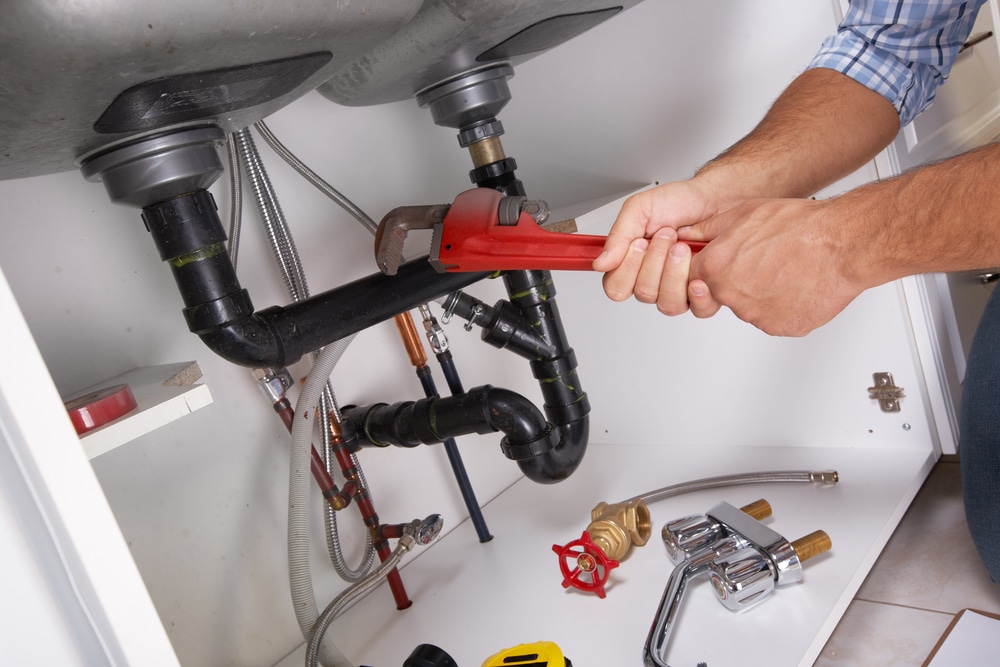 hands reaching beneath sink with plumbing tools to work on pipes